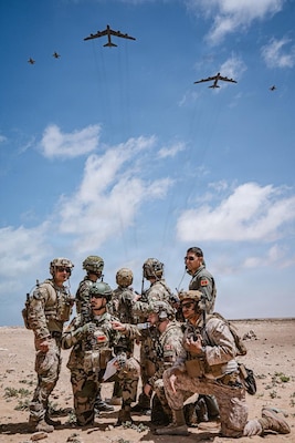 Eight men in camouflage military uniforms, helmets and sunglasses and another man in a flight suit, pose for a photo in a desert location, as five large military aircraft fly overhead.