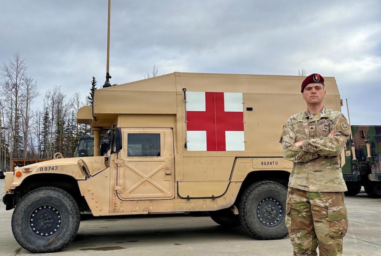 A man in a military camouflage uniform poses for a photo outside with his arms crossed, standing in front of a military medical vehicle.