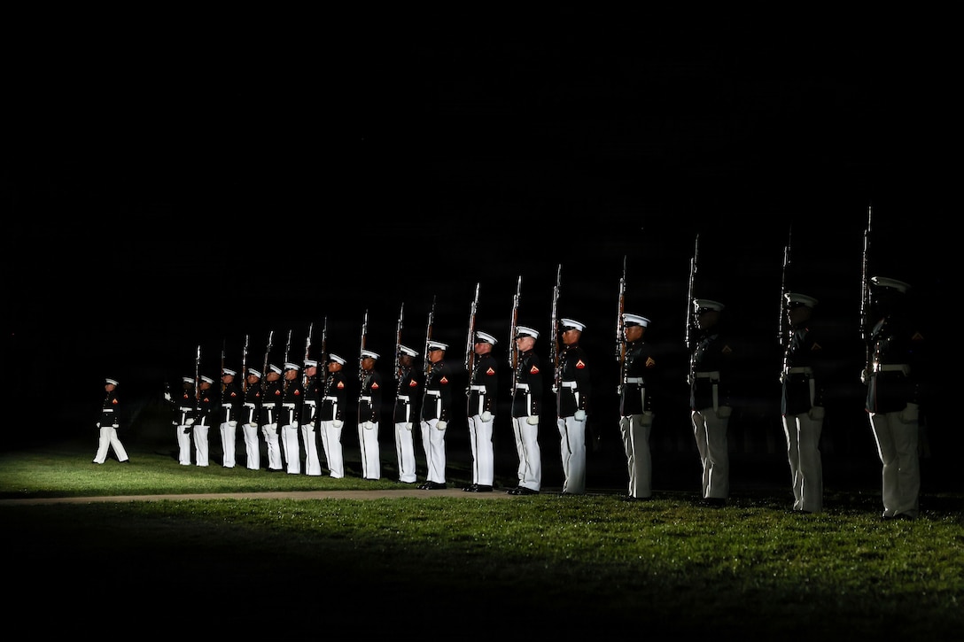 A Marine steps forward as dozens of fellow Marines in ceremonial dress stand in formation while holding rifles on a dark field illuminated by a spotlight.