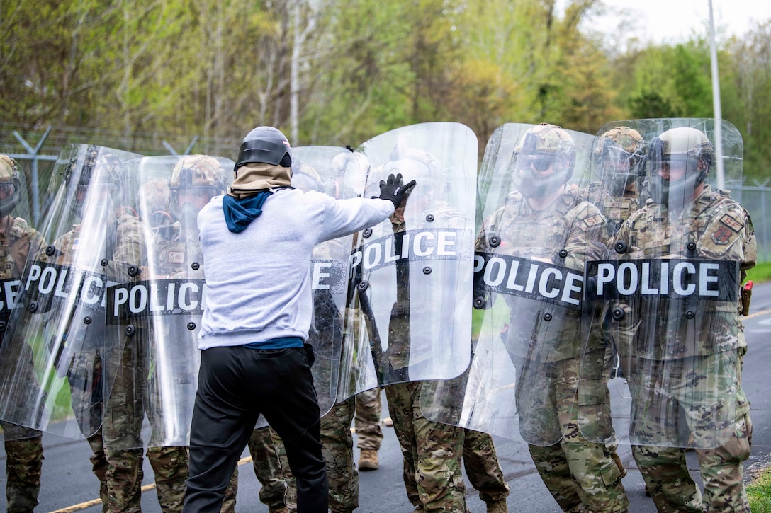 Airmen standing in formation in tactical gear hold transparent police shields as a person in civilian clothes presses against them.
