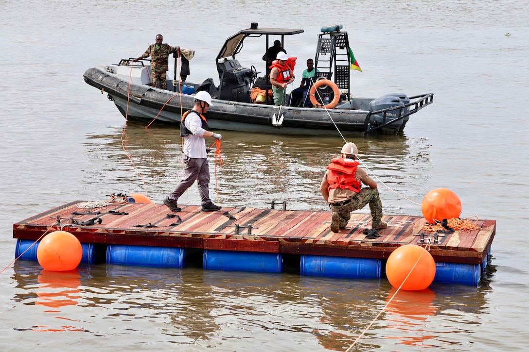 Two people stand on a floating dock while pulling a line connected to a small boat during the day.