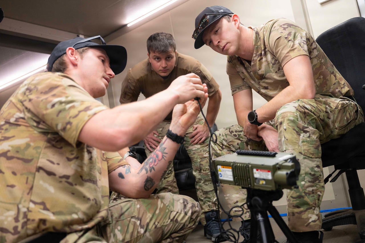 Three men wearing camouflage military uniforms familiarize themselves with a hand-held laser device.