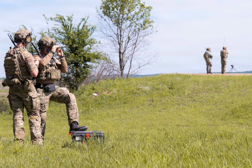 Two people wearing camouflage military uniforms and combat gear stand at the bottom of a small, grass-covered hill, with one looking through binoculars. There are two people in similar attire standing at the top of the hill.