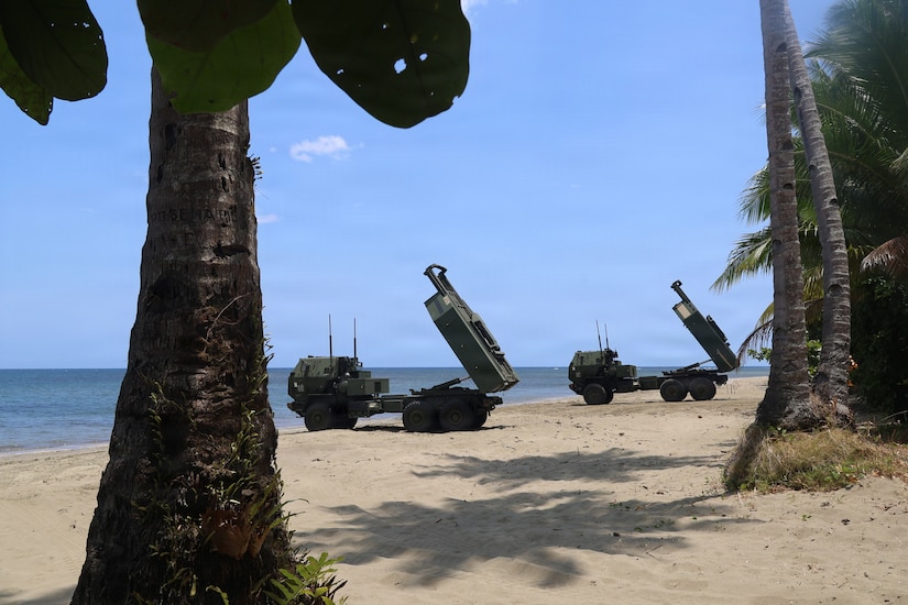 Two military vehicles are parked on a tropical beach.