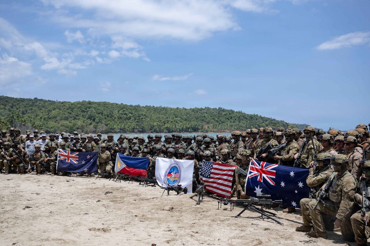 A large number of military personnel dressed in various camouflaged uniforms pose for a photo with flags from various countries.