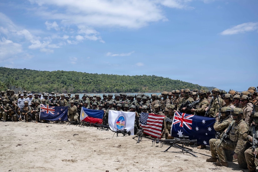 A large number of military personnel dressed in various camouflaged uniforms pose for a photo with flags from various countries.