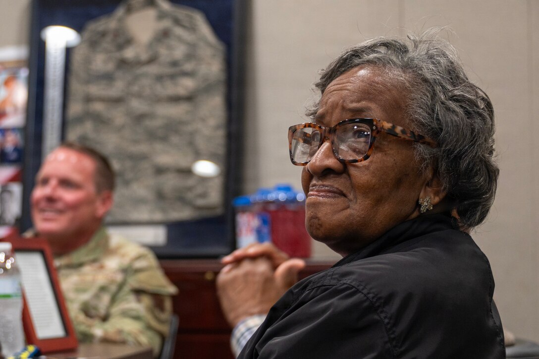 An elderly woman listens to a speaker in a conference room while a uniformed service member adjacent to her.