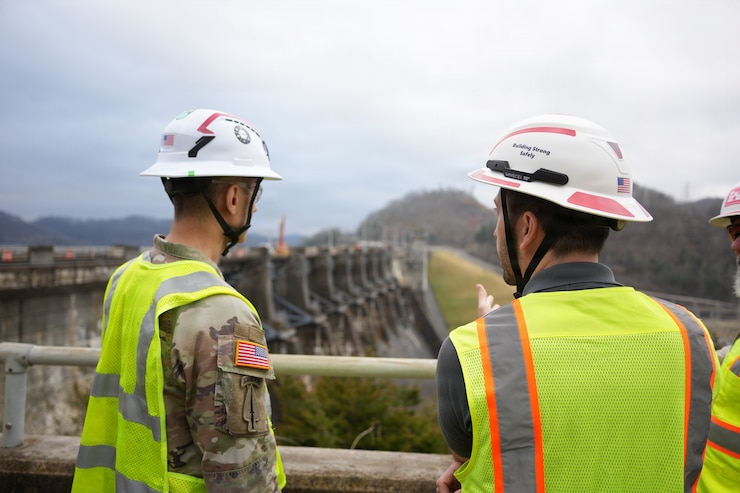 Photo of two USACE personnel from behind, wearing hard hats and safety vests, overlooking a dam.