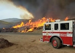 The cab of a fire truck is in the foreground while a wildfire burns on a grassy landscape of hills.