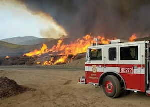 The cab of a fire truck is in the foreground while a wildfire burns on a grassy landscape of hills.