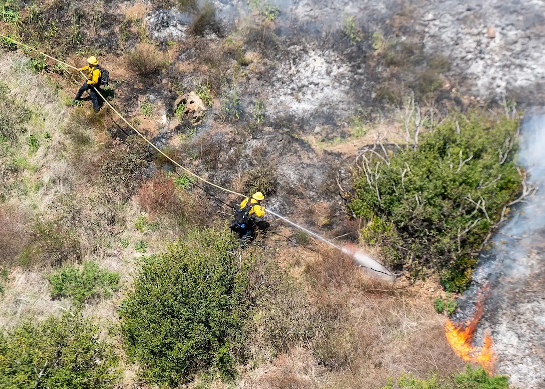 Two fire fighters are seen from the air attacking a fire with a hose and water in what looks like tall grass and scrubby trees.