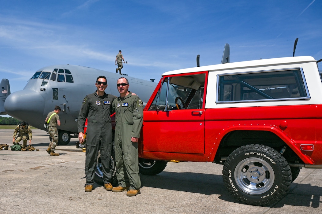 Two men in uniform lean against a red car with a C-130J Super Hercules aircraft in the background on a flightline.