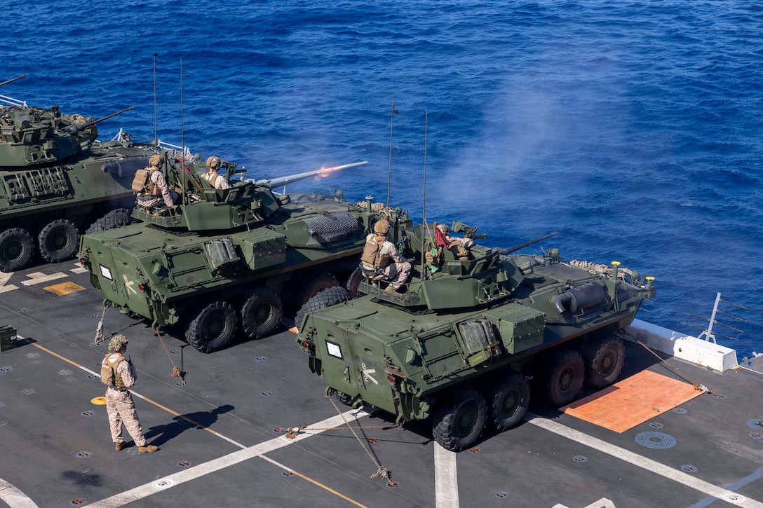 Marines sit on tanks and fire from the deck of a ship, with the ocean in the background.