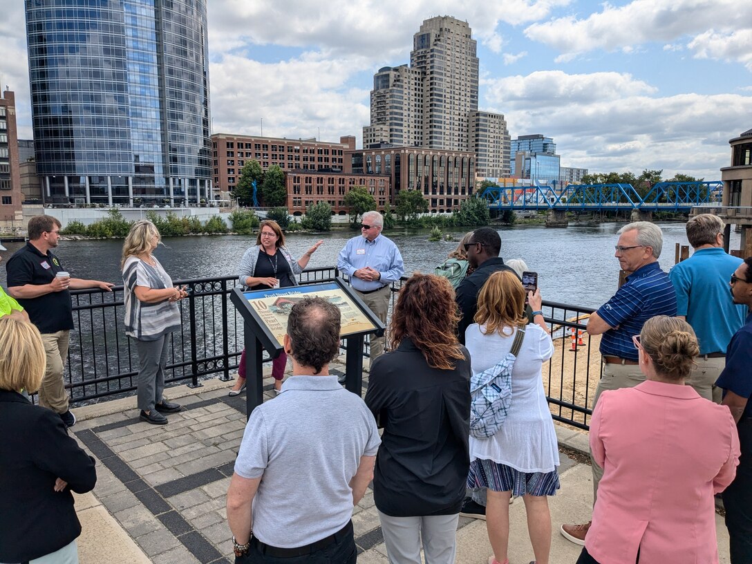 Group of approximately 15 people gathered around interpretive sign on riverwalk, Grand River and blue bridge visible behind them, downtown Grand Rapids skyline in background