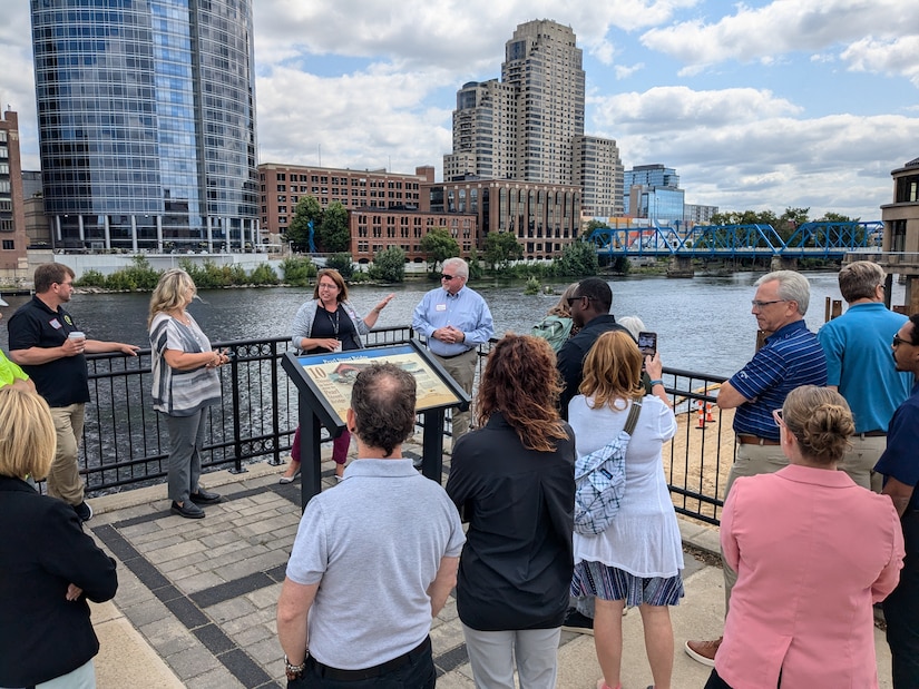 Group of approximately 15 people gathered around interpretive sign on riverwalk, Grand River and blue bridge visible behind them, downtown Grand Rapids skyline in background