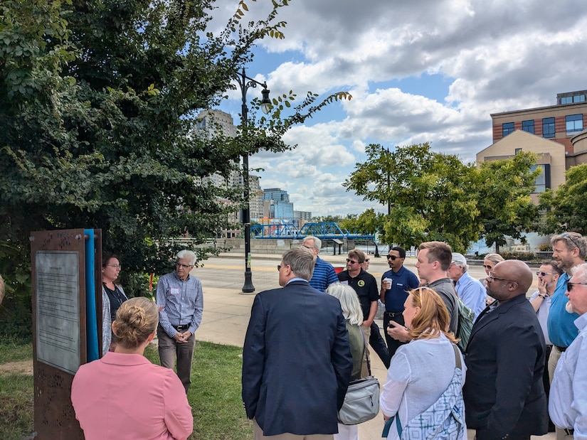 Group of approximately 20 people gathered on riverwalk listening to presentation, informational kiosk visible on left, blue bridge and downtown buildings in background under partly cloudy sky