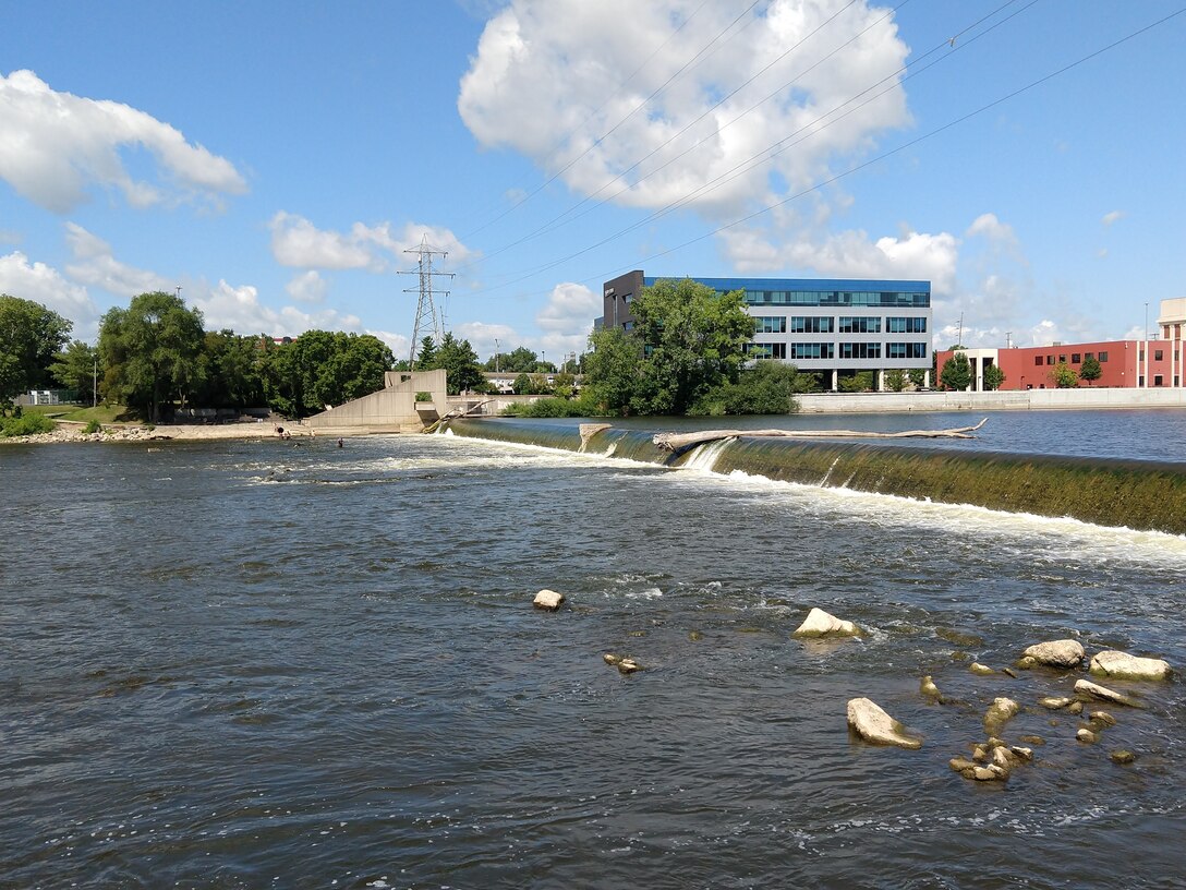 View of 6th Street Dam with water flowing over concrete structure in multiple sections, rocks visible in foreground, downtown buildings and power lines in background