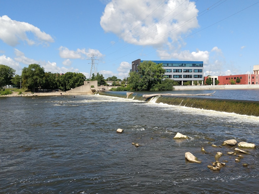 View of 6th Street Dam with water flowing over concrete structure in multiple sections, rocks visible in foreground, downtown buildings and power lines in background