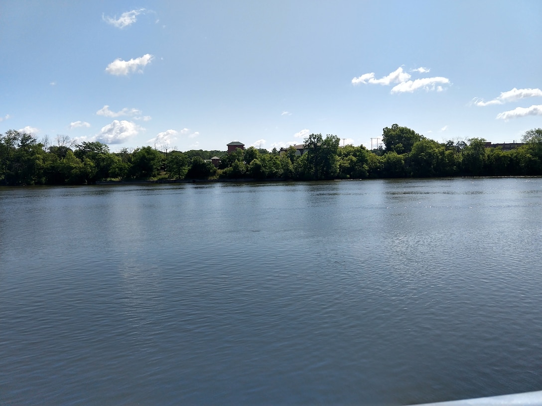 View of calm Grand River with heavily wooded shoreline, water tower visible in distance, blue sky with scattered clouds, taken from water level