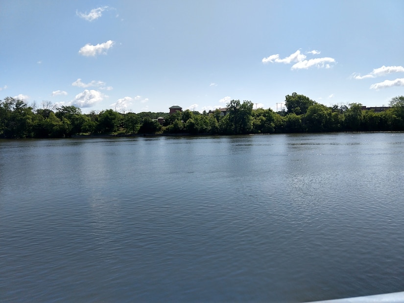 View of calm Grand River with heavily wooded shoreline, water tower visible in distance, blue sky with scattered clouds, taken from water level
