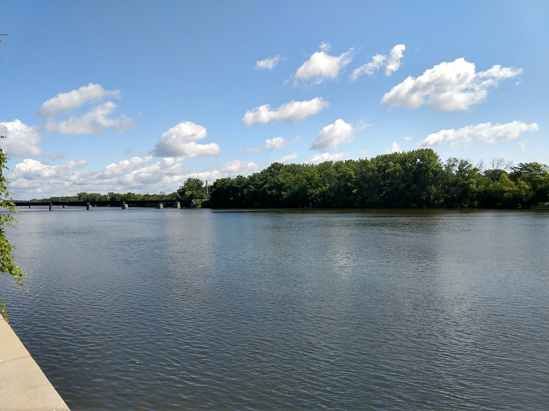 Wide view of calm Grand River with wooded shoreline, bridge visible in distance, blue sky with white clouds, concrete riverbank in foreground