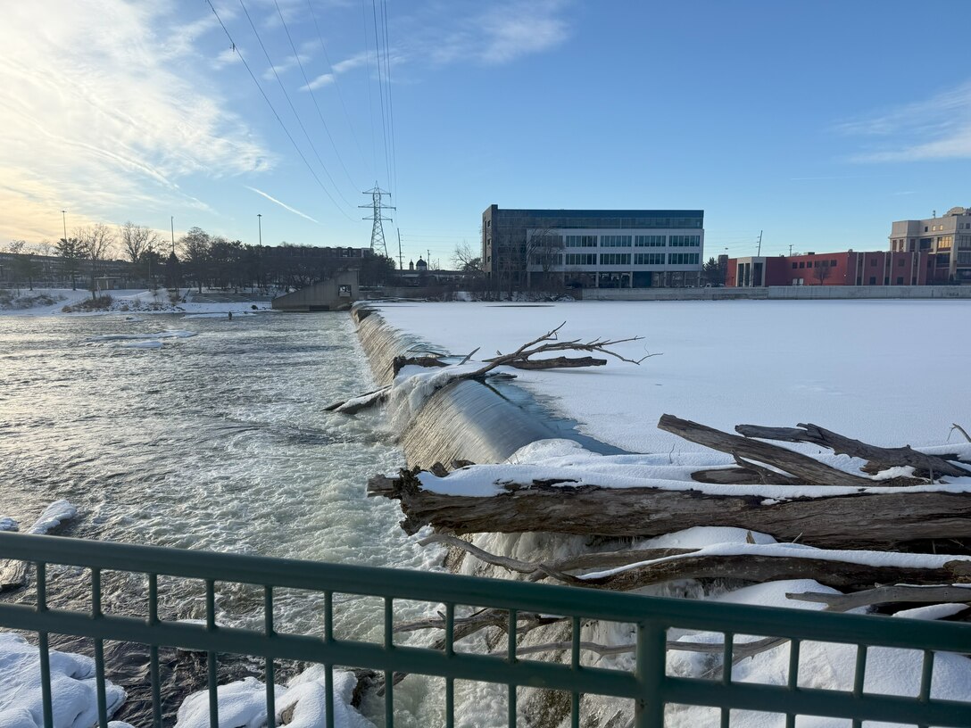 View of 6th Street Dam with water flowing over structure, logs and debris visible, snow and ice on riverbanks, downtown buildings in background