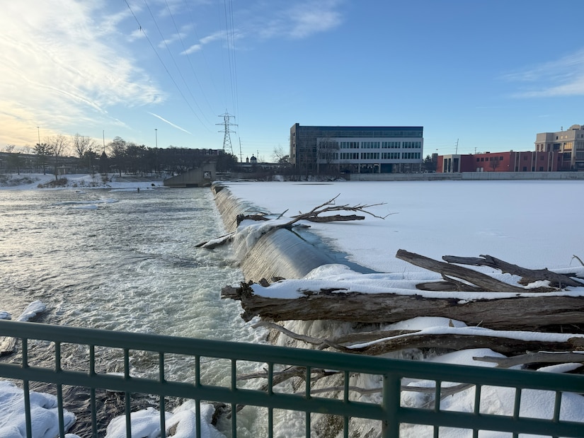 View of 6th Street Dam with water flowing over structure, logs and debris visible, snow and ice on riverbanks, downtown buildings in background
