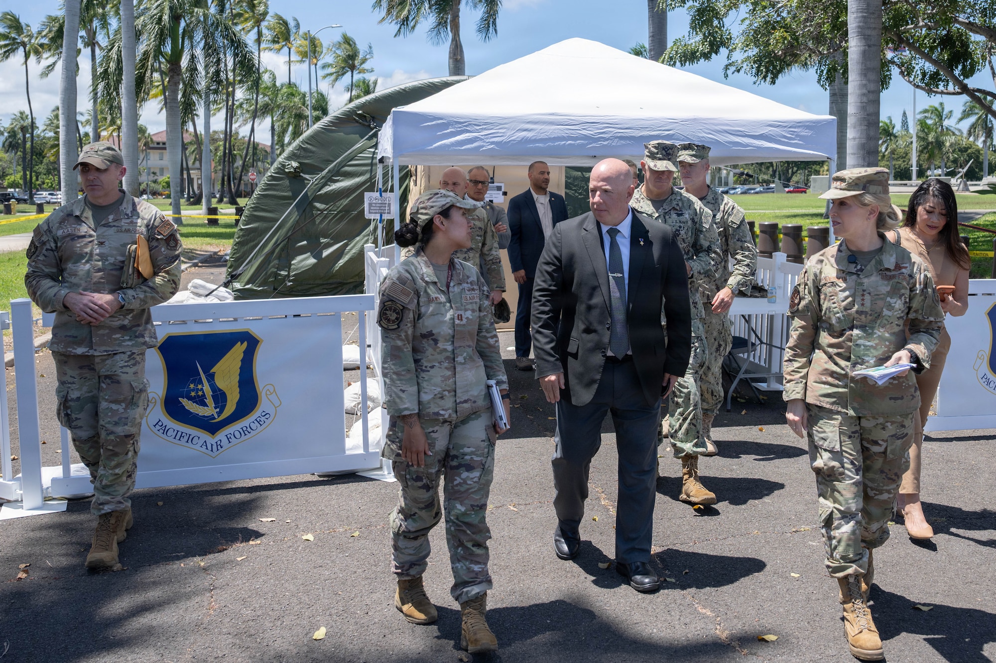 A group of USAF military members walk while looking and speaking with a man in a suit in the center. They are outside and behind then is a tent gazebo.