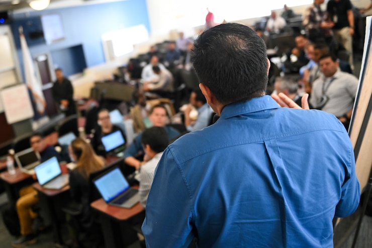 A man, from behind, wearing a blue shirt and standing in front of a class of students on their laptops.