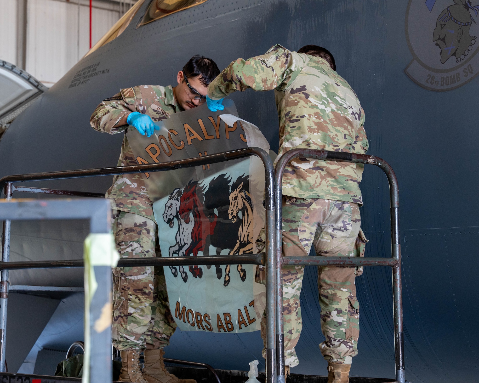 From left, U.S. Air Force Airman 1st Class Giovanni Vasquez and Senior Airman Colton Decker, both 7th Equipment Maintenance Squadron aircraft structural maintenance journeymen, prepare the “Apocalypse II” nose art for application at Dyess Air Force Base, Texas, April 27, 2026. The decal application marked a major milestone in returning the recently regenerated B-1B Lancer to operational status to serve as the 7th Bomb Wing's new flagship. (U.S. Air Force photo by Airman 1st Class William Neal)