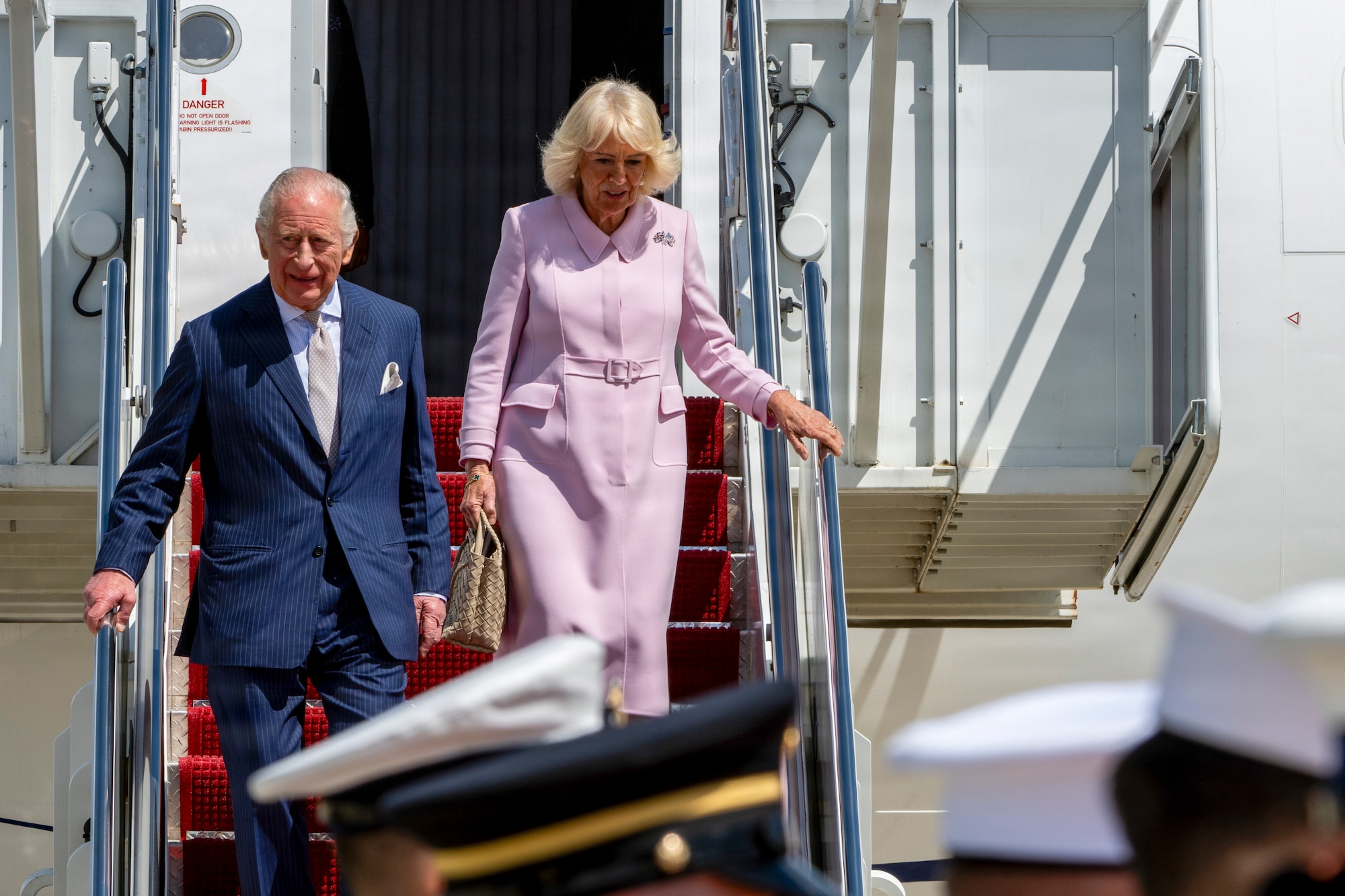 Their Majesties of the United Kingdom of Great Britain and Northern Ireland, King Charles III and Queen Camilla, descend the stairs of a United Kingdom aircraft during a military arrival ceremony.