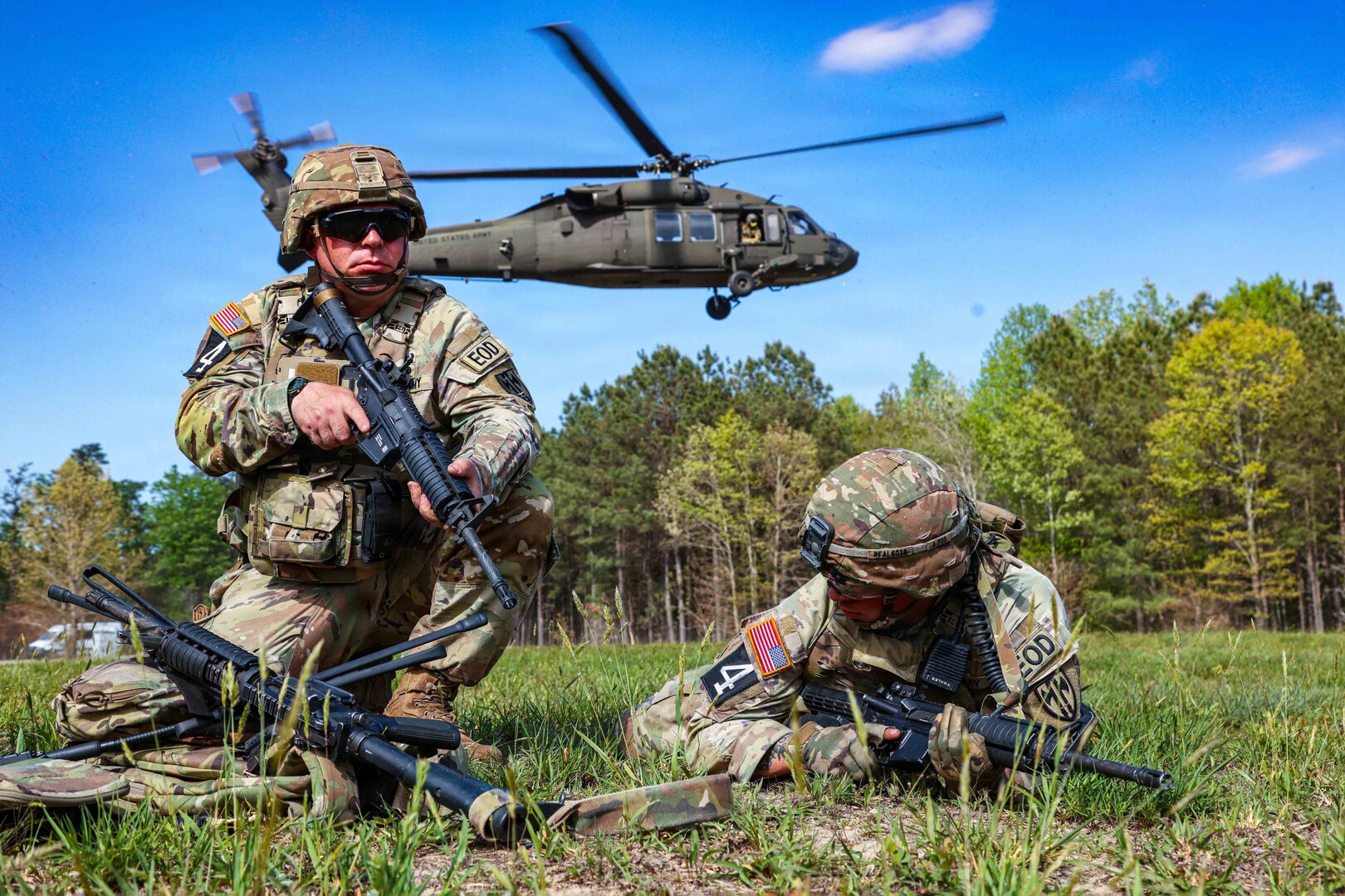 One soldier kneels as the other lies on the ground, both with weapons, as a helicopter hovers in the background over a wooded area during the day.