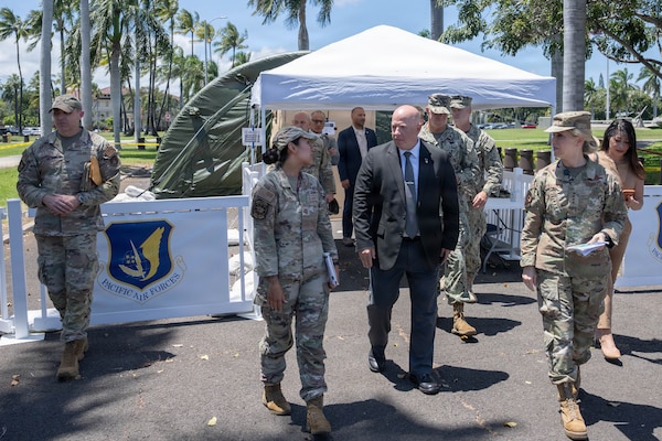 A group of USAF military members walk while looking and speaking with a man in a suit in the center. They are outside and behind then is a tent gazebo.