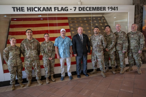 A group of USAF military members with a man in a suit and another man in casual attire standing in the center, pose for a photo in front of the old restore flag, Ol' Glory and the words above on the wall "THE HICKAM FLAG - 7 DECEMBER 1941."