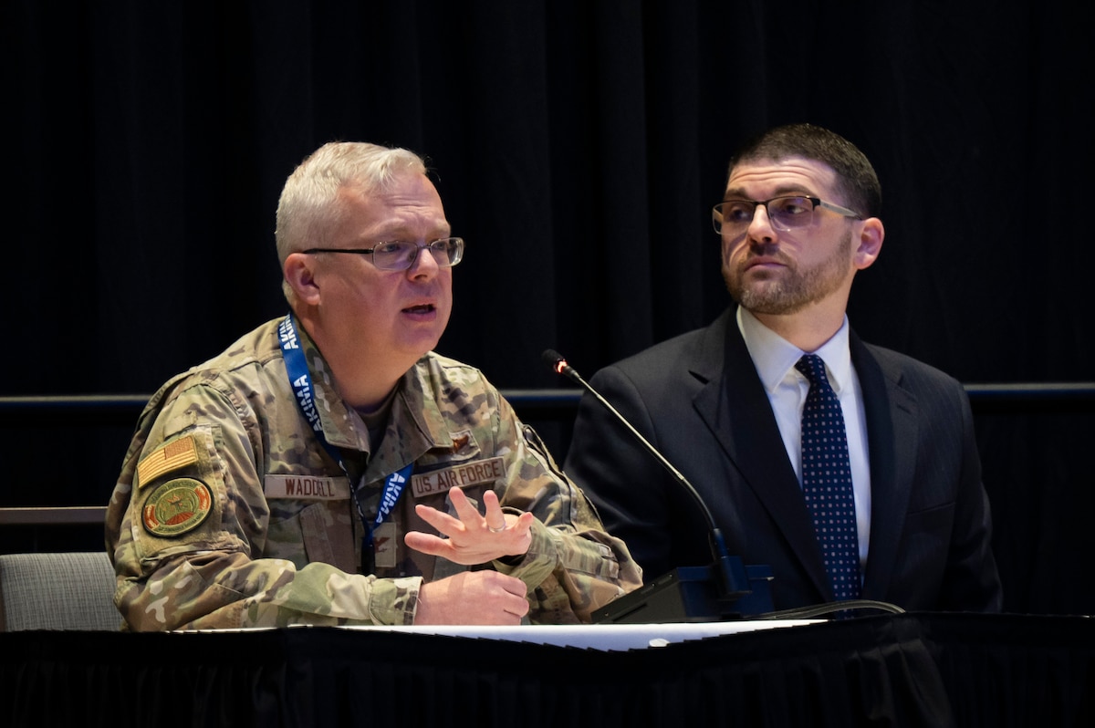 Two men sit at table with microphone. Man in Air Force camo speaks into mic.