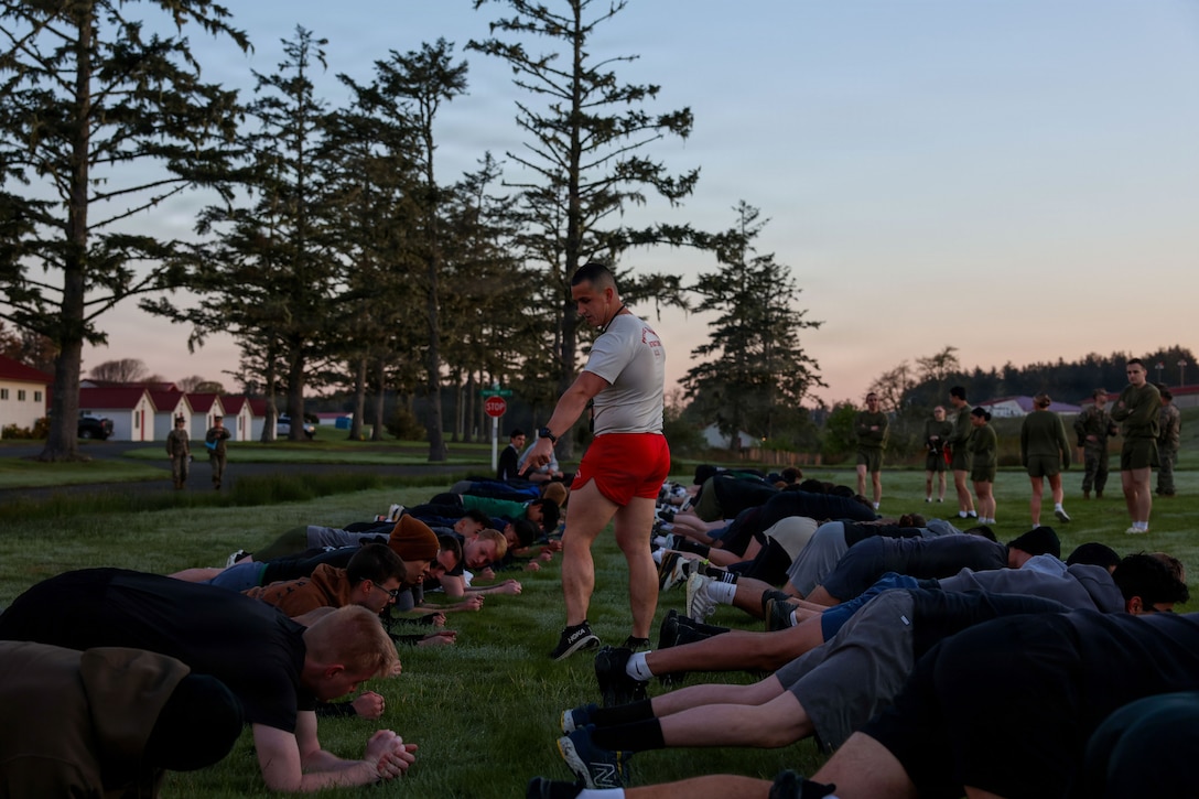 U.S. Marine Corps Sgt. Ethan Carr, a physical training instructor with Officer Candidate School, oversees officer candidates performing planks during a physical fitness test at a Mini-Officer Candidate School event at Camp Rilea Armed Forces Training Center in Warrenton, Oregon, April 25, 2026. Mini-OCS provides applicants and candidates a glimpse into Officer Candidate School by placing them through an intense training environment designed to test their endurance, resiliency, and courage. (U.S. Marine Corps photo by Lance Cpl. Monserrath Mora)