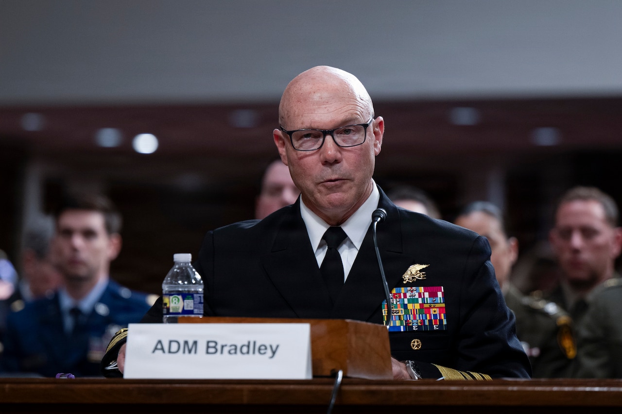 A Navy admiral in a formal military uniform sits at a table and speaks into a microphone by a place card that reads, "ADM Bradley."