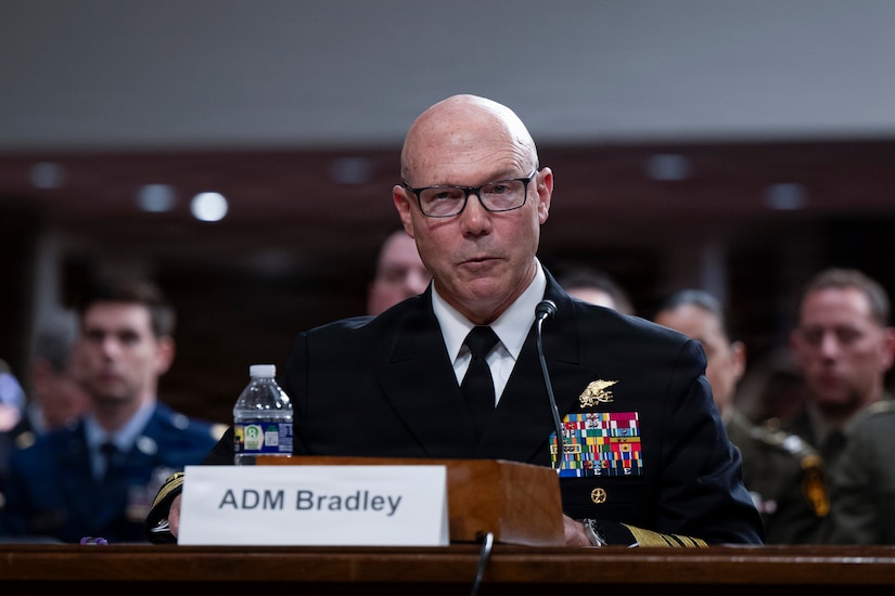 A Navy admiral in a formal military uniform sits at a table and speaks into a microphone by a place card that reads, "ADM Bradley."