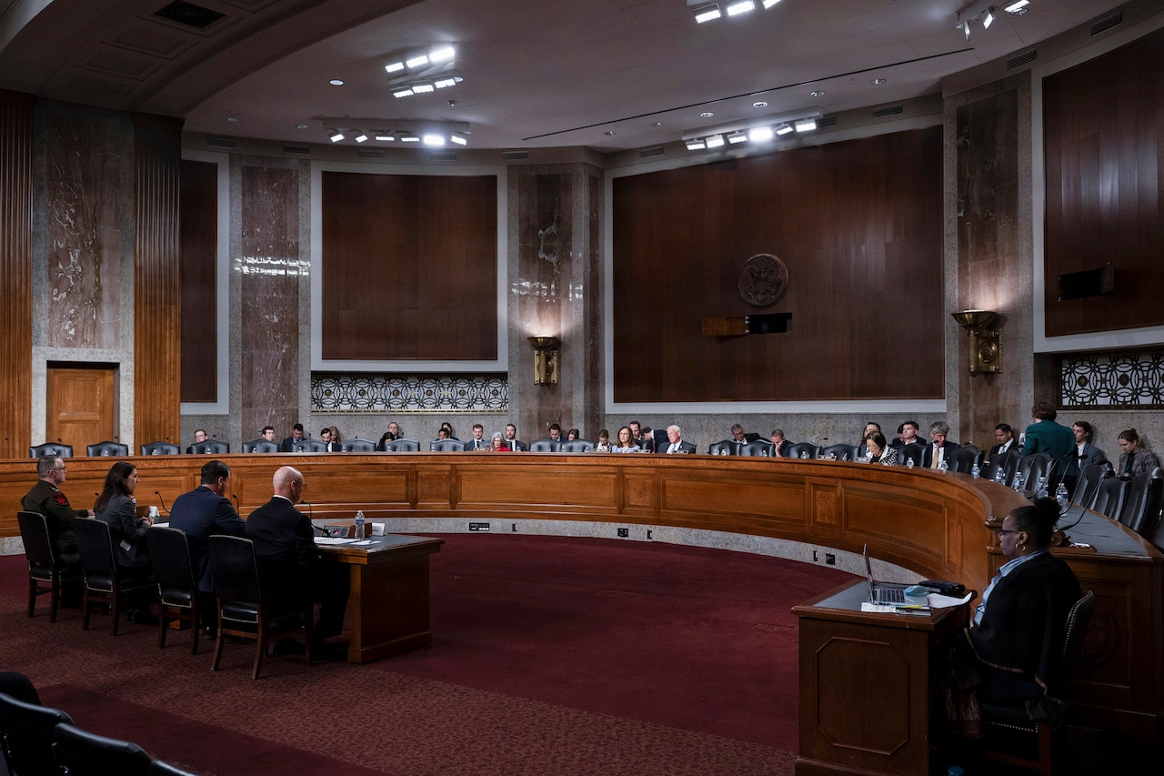 In a large, stately room with high ceilings, lawmakers sit on an elevated, circular dais, looking down toward four individuals who are sitting at a small table in front of them.