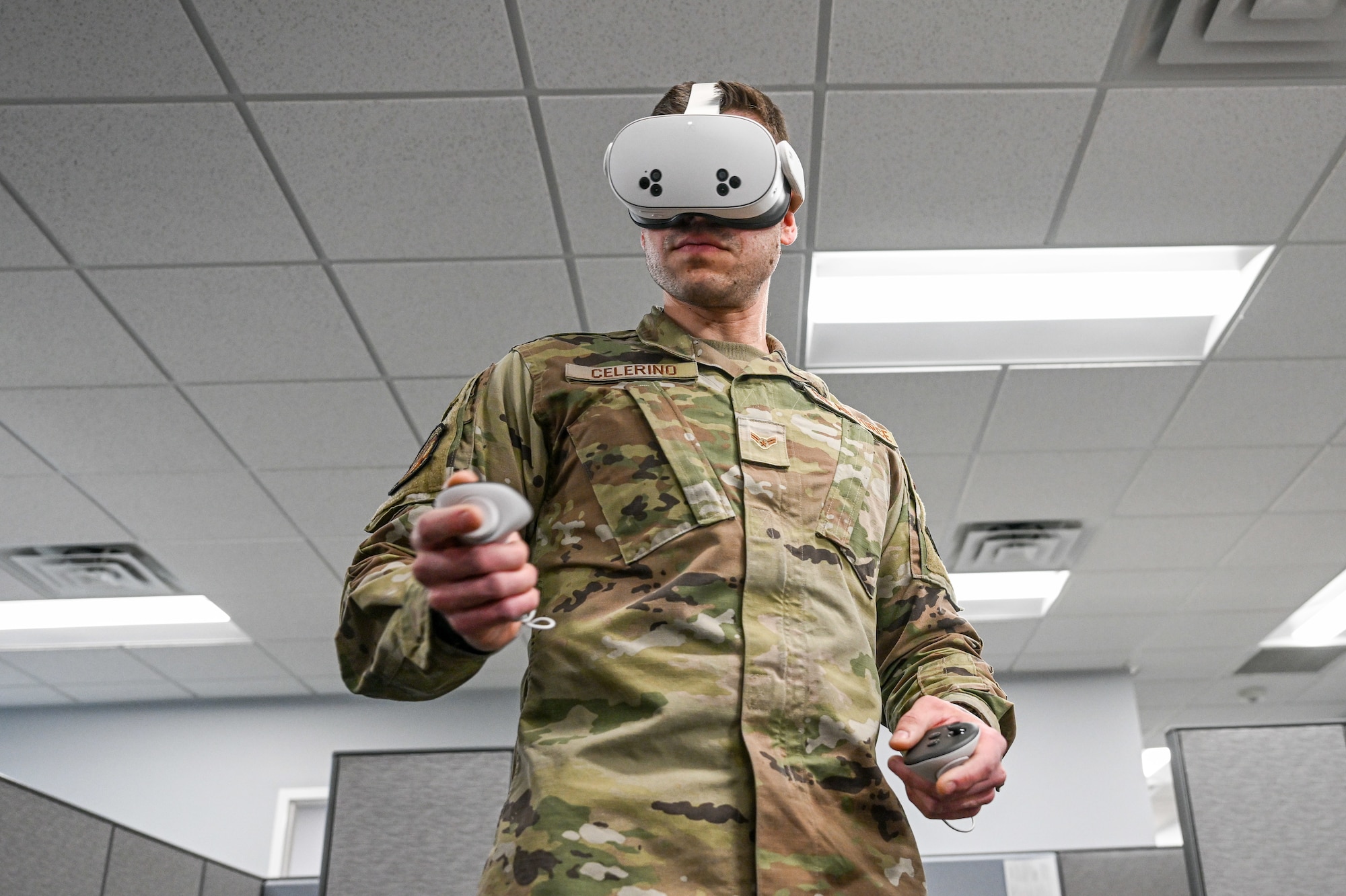 Airman in camouflage uniform using a virtual reality headset and handheld controllers in an indoor office setting.