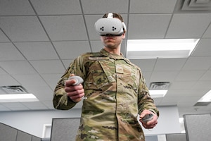 Airman in camouflage uniform using a virtual reality headset and handheld controllers in an indoor office setting.