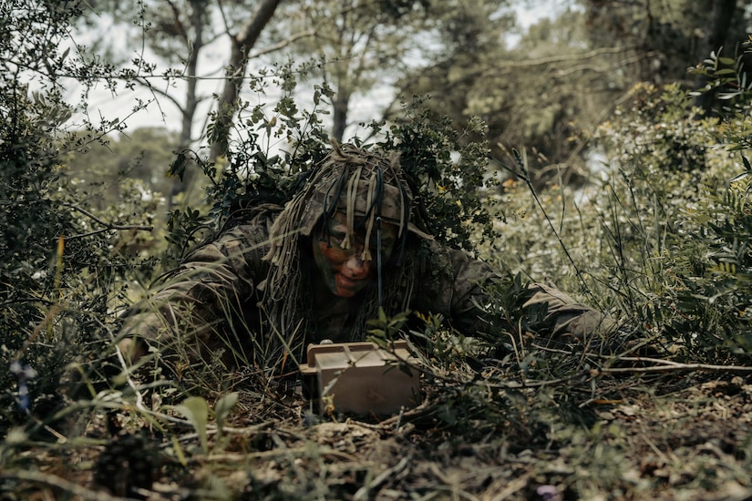 A man in a camouflage military uniform and face paint lies on the ground in the woods, looking at a piece of surveillance equipment.
