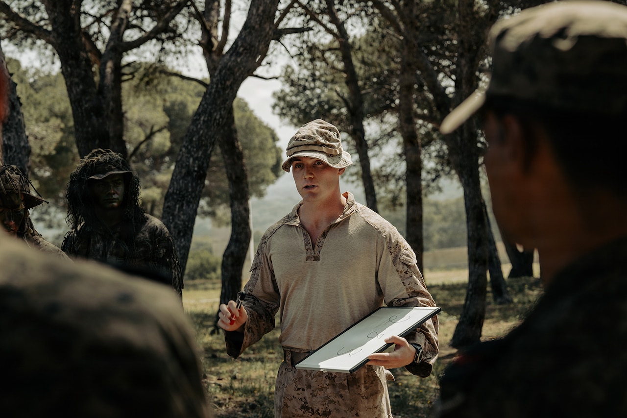 A man in a camouflage military uniform speaks to a group of people in similar attire while standing in the woods holding a dry-erase board.
