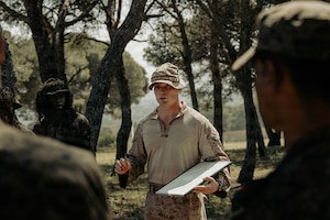 A man in a camouflage military uniform speaks to a group of people in similar attire while standing in the woods holding a dry-erase board.