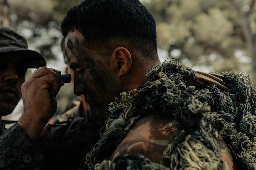 A man in a camouflage military uniform applies face paint to another man in similar attire in the woods.
