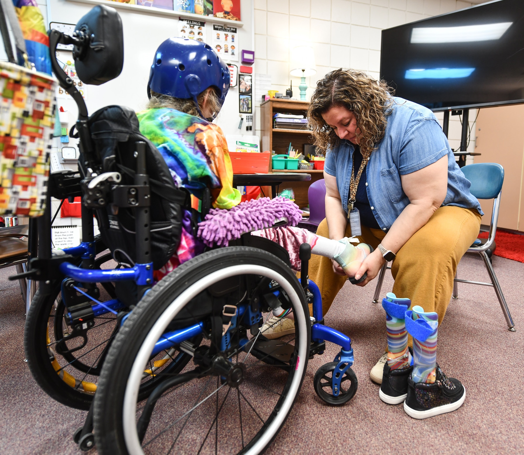 Ms. Rhonda Stacey helps Hannah switch her shoes out after recess at Leonard Lawrence Elementary school on April 22, 2026. Hannah needs special equipment to function in her day-to-day activities—a wheelchair to get around, special clothes and shoes to play in, and a helmet in case she has a seizure. (U.S. Air Force photo by Chad Watkins)