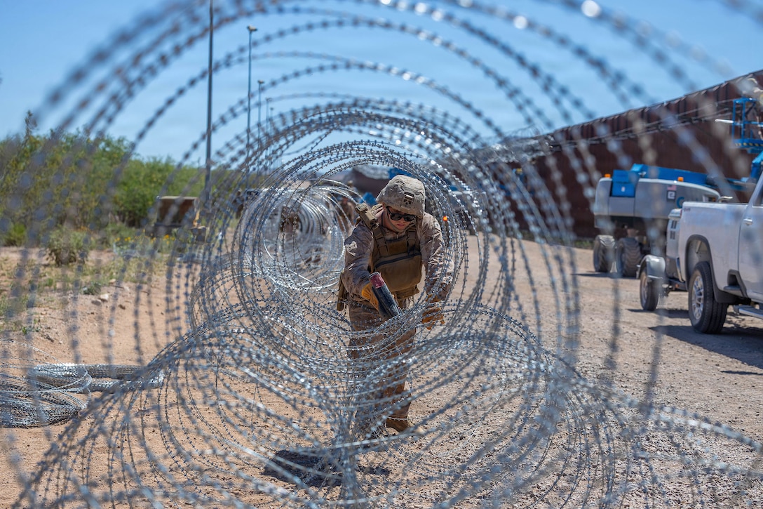 A Marine uses a tie gun on coiled wire sitting on the ground outdoors near vehicles parked by a tall wall.