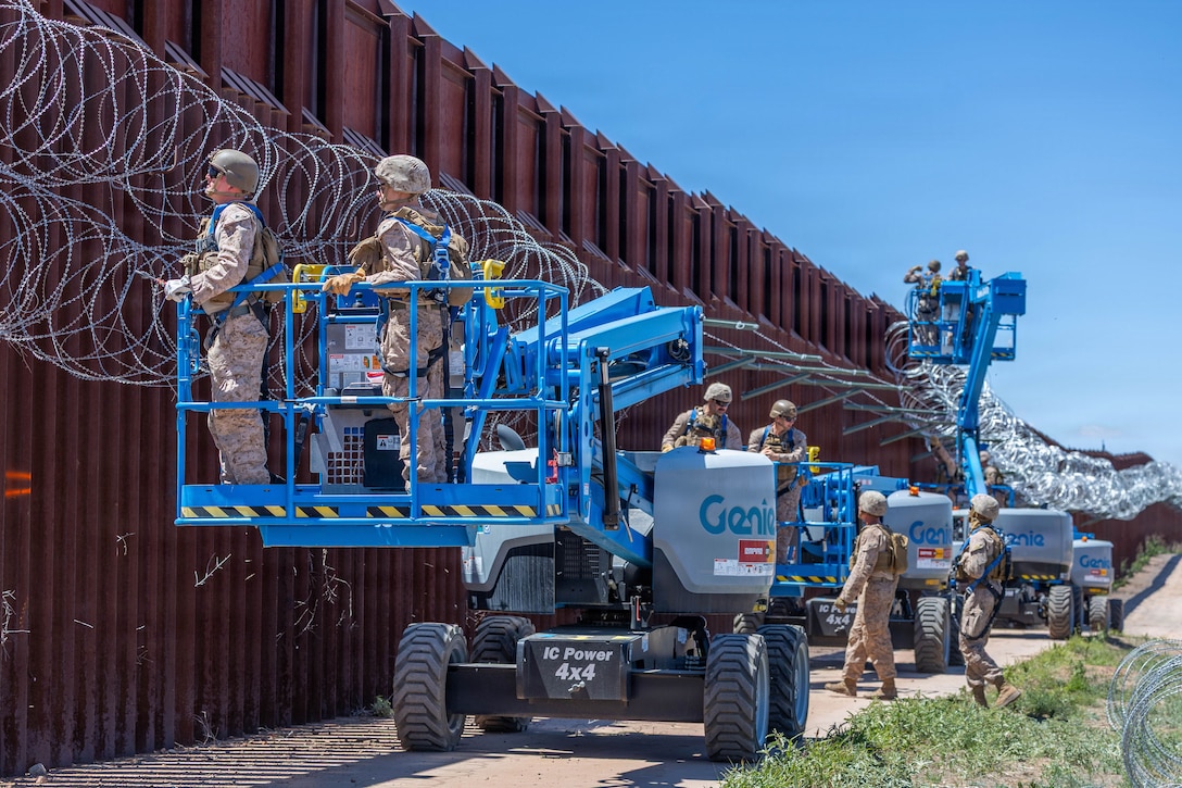 Marines stand on raised platforms attached to vehicles and handle concertina wire attached to an outdoor wall.