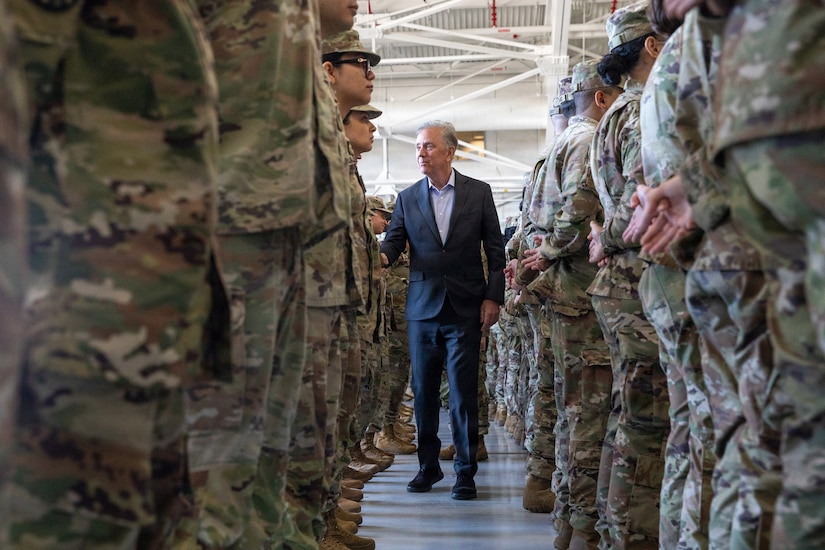 A man in a blue suit walks between a formation of men and women dressed in camouflage military uniforms.