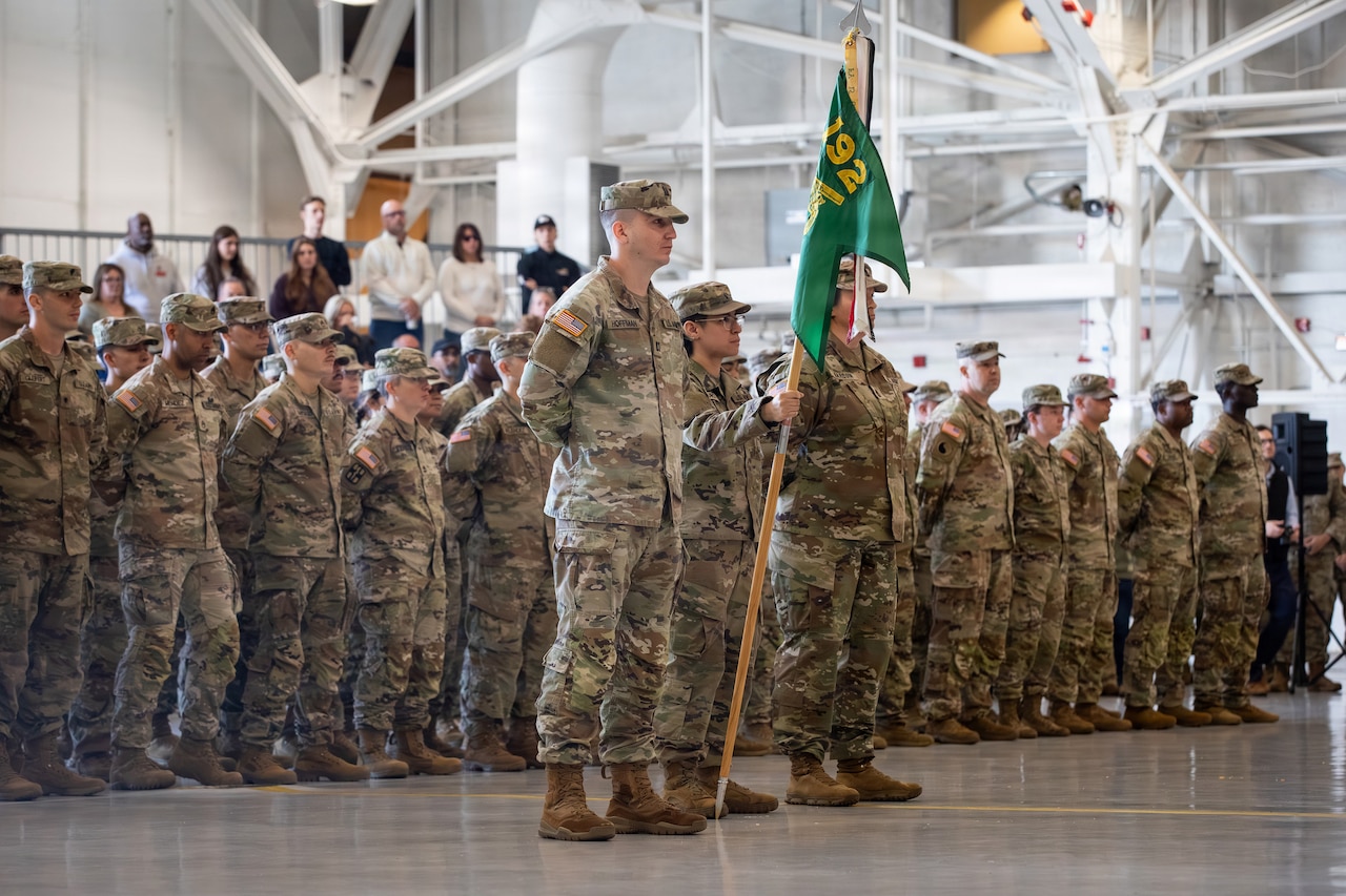 Men and women dressed in camouflage military uniforms stand in formation. A man at the front of the formation holds a wooden pole with a green flag attached to the top of it while two people in similar attire stand next to him.